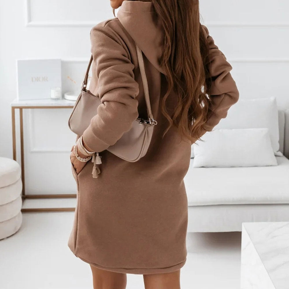 Frau in braunem Kapuzenkleid mit beiger Handtasche, modernes Wohnzimmer, gemütlicher Stil.