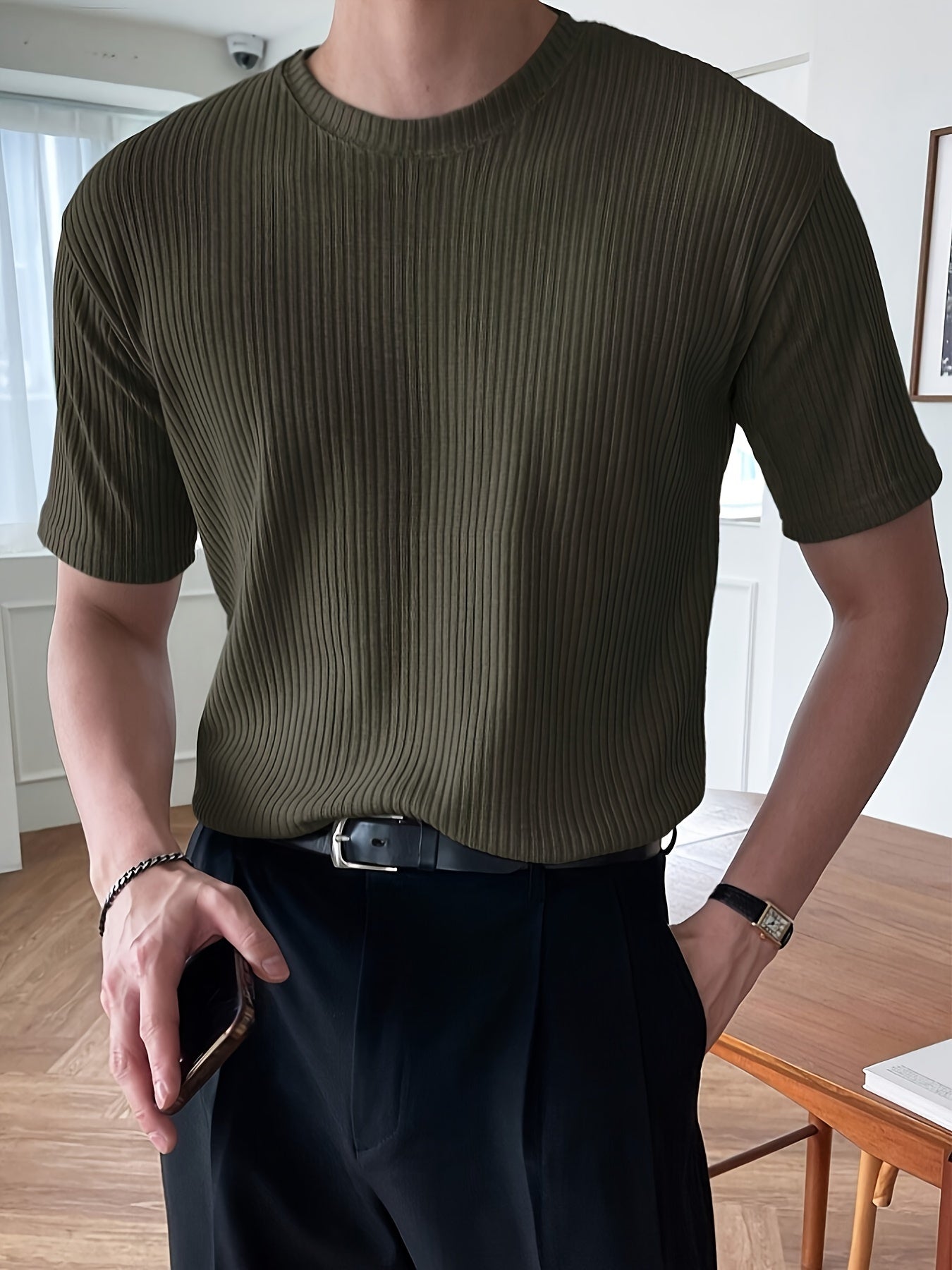 Mann in olivgrünem, geripptem T-Shirt, schwarze Hose, Armbanduhr, Smartphone in der Hand.