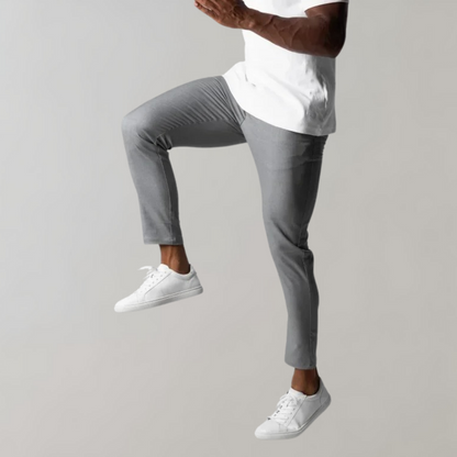 Mann in grauer Hose und weißen Sneakers, springend, vor grauem Hintergrund.