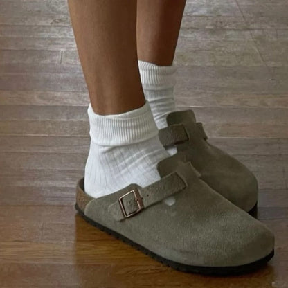 Graue Wildleder-Clogs mit Schnalle, getragen mit weißen Socken auf Holzboden.